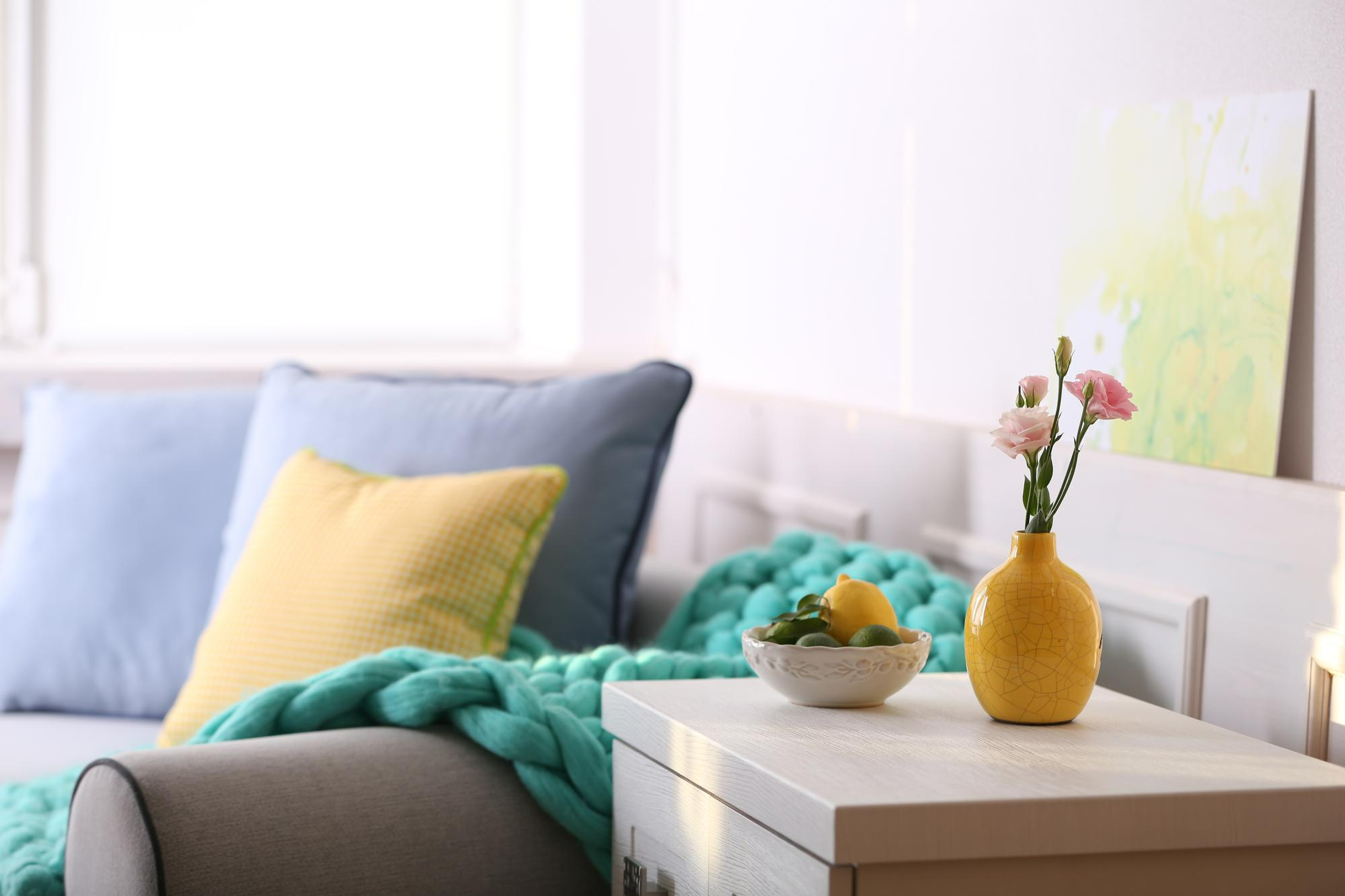 grey couch with yellow throw pillow and blue blanket and pops of color and fresh flower accents on table creating a Spring aesthetic for your apartment
